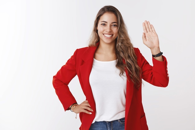 Atractiva joven europea de pelo rizado de 25 años de aspecto amistoso, confiada, vestida con chaqueta roja, saludando con la palma levantada, gesto de bienvenida, sonriendo, saludando a los miembros del equipo, diciendo hola conociendo gente nueva