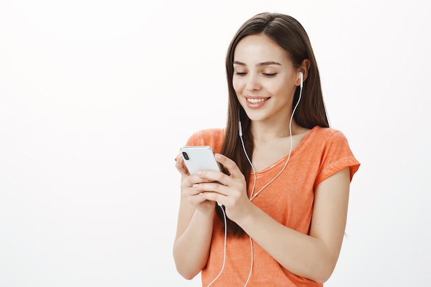 Atractiva joven morena escuchando música en auriculares y enviando mensajes de texto, usando el teléfono móvil, disfrutando de un interesante podcast