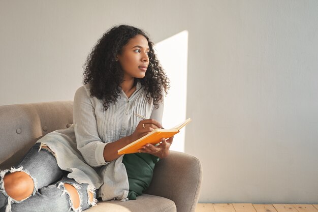 Atractiva joven mulata de piel oscura con peinado afro relajándose en el sofá en casa, con mirada pensativa y pensativa, escribiendo ideas para su propio proyecto de inicio, usando bolígrafo y cuaderno