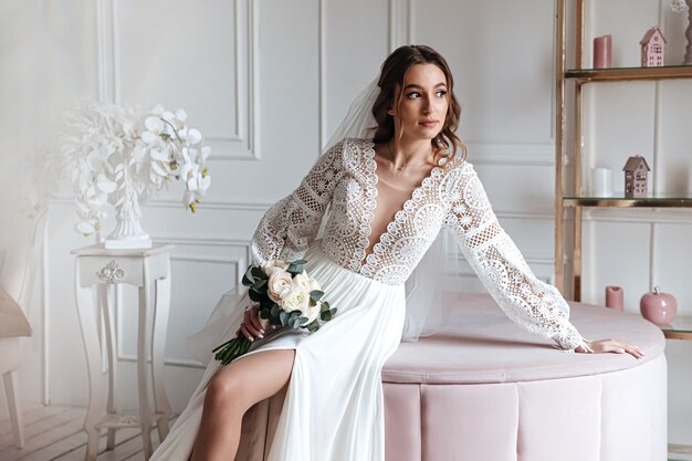 Una atractiva joven novia con un hermoso vestido de novia boho posando con un ramo de flores en una habitación luminosa.