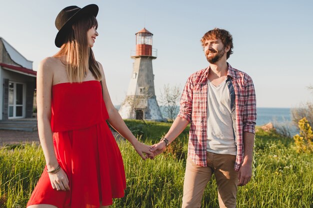 Atractiva joven pareja elegante enamorada en el campo, estilo bohemio indie hipster, vacaciones de fin de semana, traje de verano, vestido rojo, hierba verde, tomados de la mano