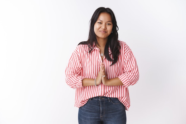 Foto gratuita atractiva joven polinesia de 20 años en blusa a rayas, presione las palmas juntas en un cortés gesto de saludo diciendo namaste inclinándose para dar la bienvenida a su querido invitado sonriendo agradablemente sobre la pared blanca