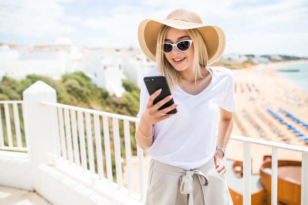 Atractiva joven rubia escribiendo en el teléfono en la terraza en la vista de la playa