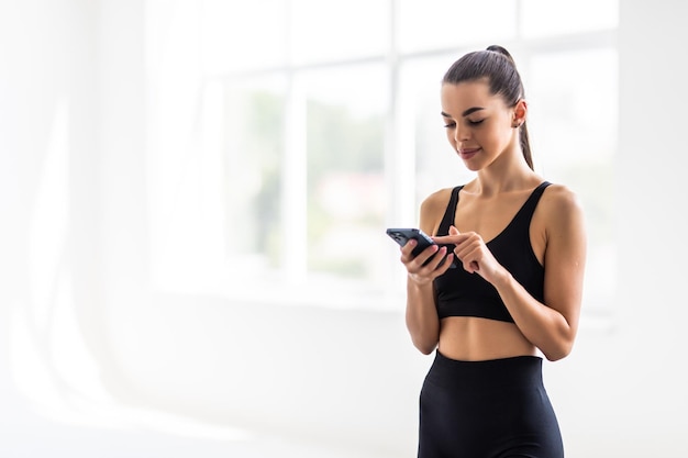 Atractiva joven saludable con una colchoneta de fitness usando un teléfono móvil en el gimnasio