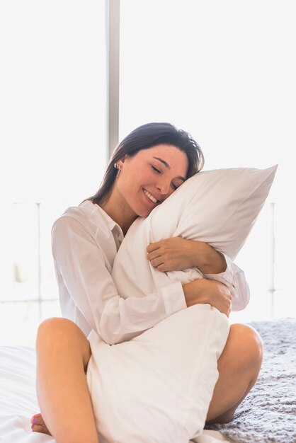Una atractiva joven sentada en la cama abrazando su almohada blanca