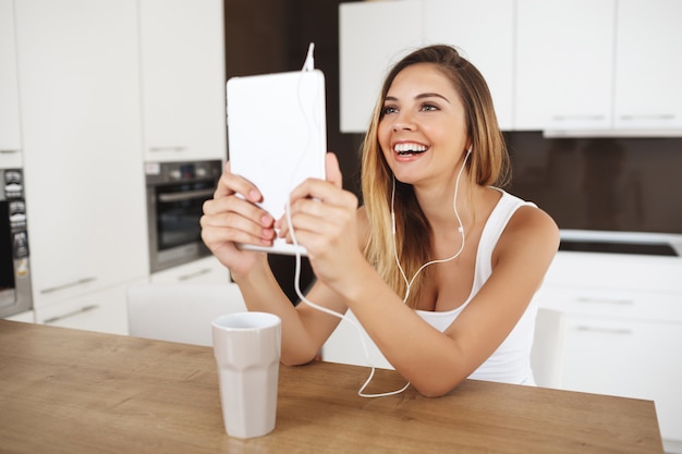 Atractiva joven sonriente sentada en la mesa con tableta y hablando con amigos a través de messenger