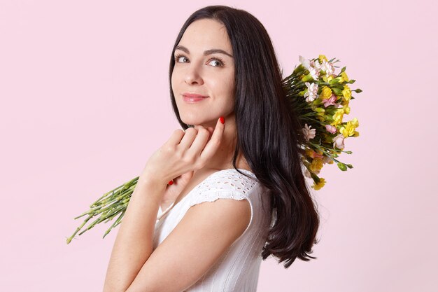 Atractiva jovencita delgada sostiene flores en su hombro, toca la barbilla con los dedos, se para de lado y mira en otra dirección