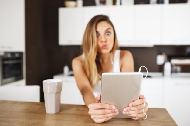 Atractiva jovencita sentada en la mesa y haciendo selfie divertido