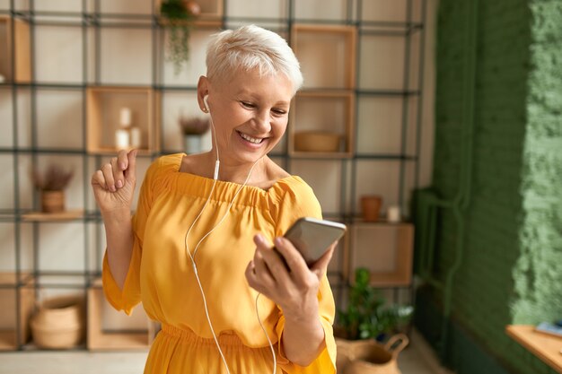 Atractiva jubilada alegre en vestido amarillo con teléfono móvil, escuchando música en auriculares, bailando, con expresión facial alegre feliz