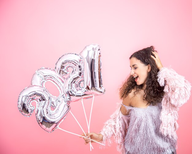 Atractiva linda chica morena con pelo rizado vestida de forma festiva posando sobre un fondo rosa con globos plateados en las manos para el concepto de año nuevo