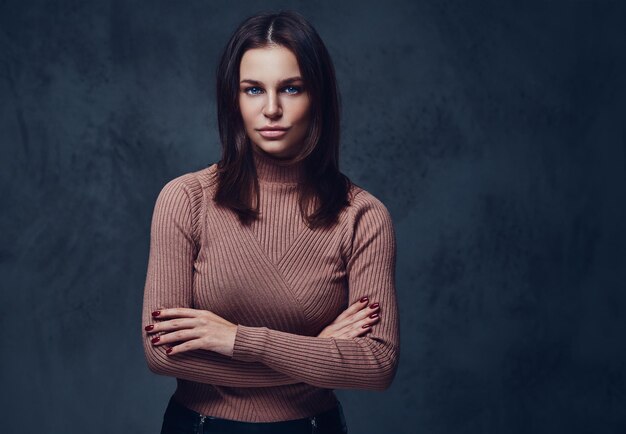 Una atractiva morena vestida con una chaqueta marrón de cuello largo sobre fondo gris.