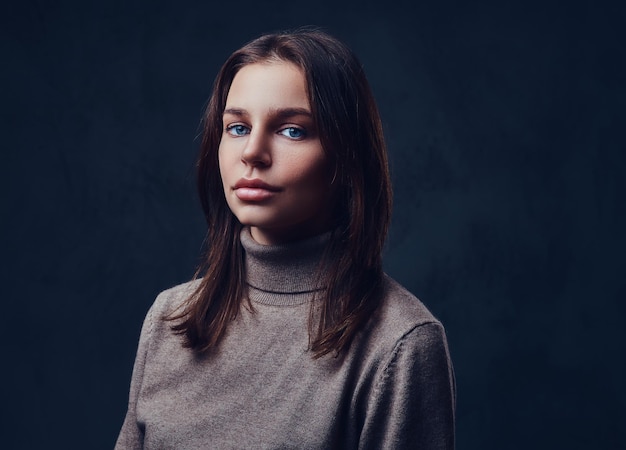 Una atractiva morena vestida con una chaqueta marrón de cuello largo sobre fondo gris.