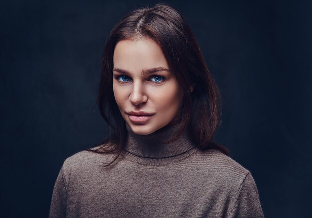 Una atractiva morena vestida con una chaqueta marrón de cuello largo sobre fondo gris.