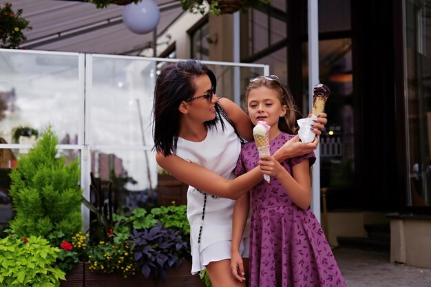 Una atractiva morena vestida con un vestido blanco y gafas de sol y una linda adolescente comiendo helado en la calle.