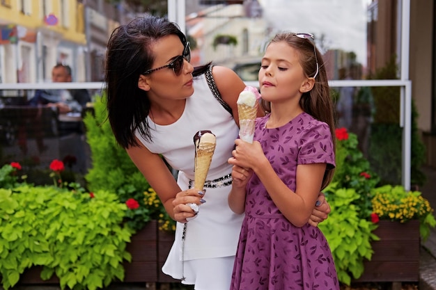 Una atractiva morena vestida con un vestido blanco y gafas de sol y una linda adolescente comiendo helado en la calle.