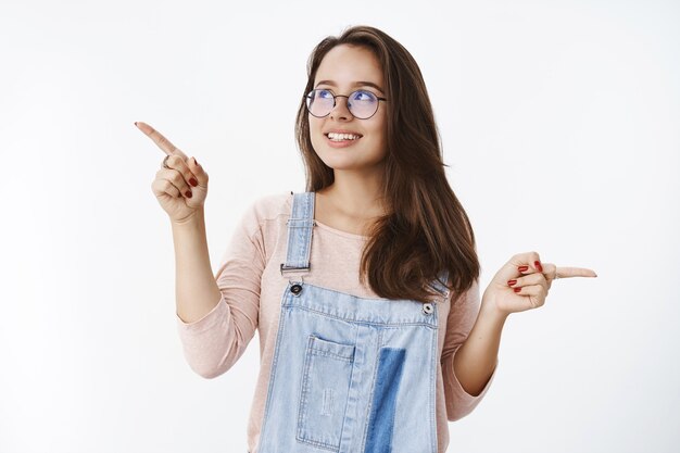 Atractiva mujer de 20 años con gafas y peto de mezclilla recogiendo producto con ojos soñadores y amplia sonrisa mirando a la esquina superior izquierda apuntando hacia los lados haciendo elección teniendo muchas variantes sobre la pared gris.