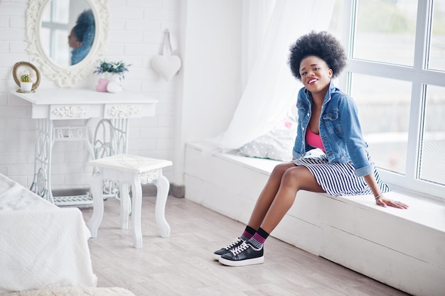 Atractiva mujer afroamericana con cabello afro en falda y chaqueta de jeans posada en la habitación blanca Modelo negro de moda