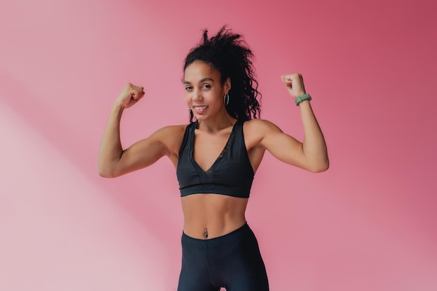 Atractiva mujer afroamericana negra en polainas negras y traje de fitness superior sobre fondo rosa aislado feliz sonriente cabello rizado estilo deportivo cuerpo atlético músculos fuertes