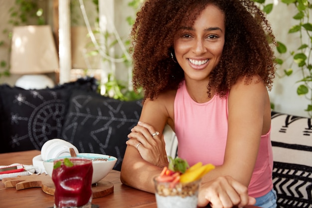 Atractiva mujer afroamericana de piel oscura vestida con camiseta rosa, se sienta con las manos cruzadas en la mesa de la cafetería, rodeada de cócteles y postre dulce, tiene expresión complacida, tiene buen descanso.