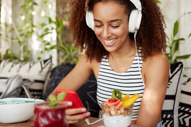 Atractiva mujer afroamericana positiva con cabello rizado usa audífonos a través de aplicaciones móviles y chats en redes, conectada a internet inalámbrico, disfruta del tiempo de recreación, lee buenas noticias