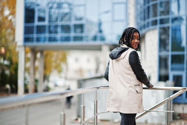 Atractiva mujer afroamericana con rastas en chaqueta posada cerca de barandillas contra un moderno edificio de varios pisos