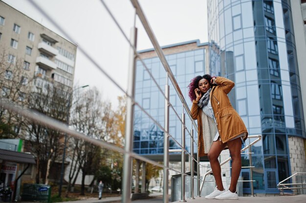 Atractiva mujer afroamericana rizada con abrigo marrón posada cerca de barandillas contra un moderno edificio de varios pisos hablando por teléfono móvil