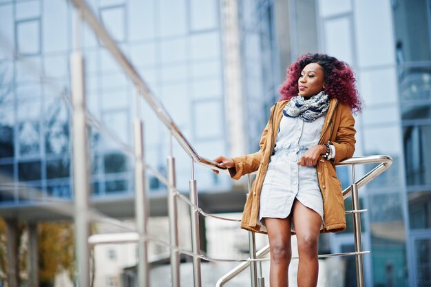 Atractiva mujer afroamericana rizada con abrigo marrón posada cerca de barandillas contra un moderno edificio de varios pisos