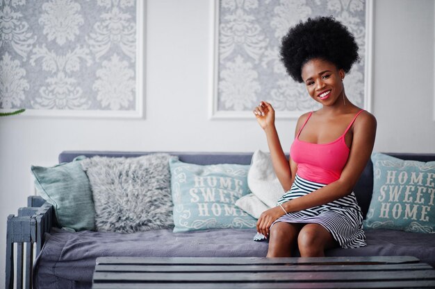 Atractiva mujer afroamericana con ropa de cabello afro en la parte superior de la falda rosa posada en la habitación blanca Modelo negro sexy de moda