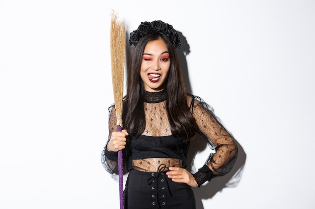 Atractiva mujer asiática descarada en traje de bruja mostrando la lengua, sosteniendo la escoba y posando sobre fondo blanco.
