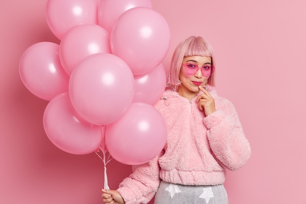 Foto gratuita atractiva mujer asiática con estilo viste peluca rosa con flecos, gafas de sol de moda, abrigo de piel con manojo de globos de helio listos para posturas de gallinas en interiores