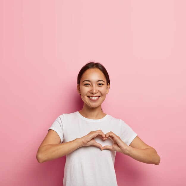 Atractiva mujer asiática hace un gesto en forma de corazón, expresa amor, dice que sea mi San Valentín, sonríe positivamente, viste un traje blanco, posa contra la pared rosa con un espacio vacío. Concepto de lenguaje corporal