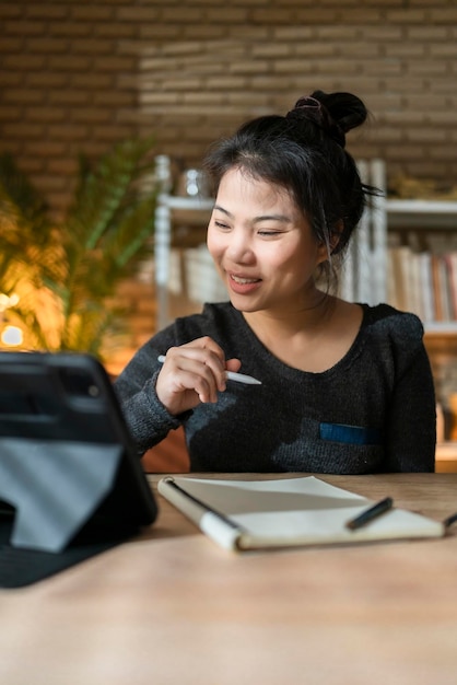 Atractiva mujer asiática inteligente usa ropa informal trabajando desde casa usando un dispositivo de tecnología de tableta mujer asiática trabaja en la oficina de estudio en casa en línea con fondo de estante de libros