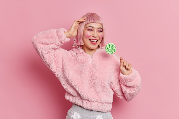 Atractiva mujer asiática joven de moda con peinado rosa sostiene un delicioso caramelo verde vestida con un abrigo de moda