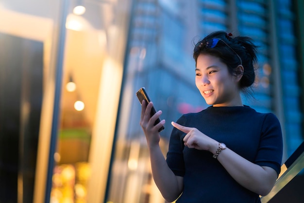 Atractiva mujer asiática mano uso teléfono inteligente tecnología comunicación con fondo de bokeh luz edificio urbano fondo concepto de ideas de negocios