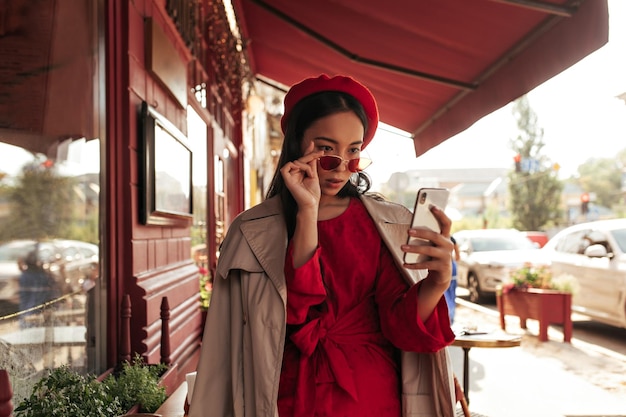 Atractiva mujer asiática morena con un elegante vestido rojo de boina y una gabardina beige se quita las gafas de sol y se toma una selfie Lady sostiene el teléfono y se apoya en la mesa en el café de la calle