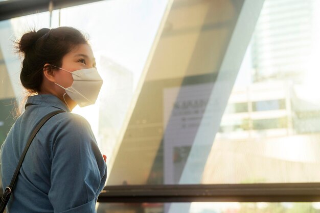 Atractiva mujer asiática de tela casual con protección de máscara facial de pie junto a grandes anteojos ventana de marco moderno con destello de luz solar momento de subconjunto nuevo concepto de atención médica de estilo de vida normal