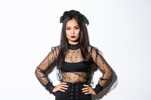 Foto gratuita atractiva mujer asiática en traje de halloween que parece decepcionada y escéptica. mujer en vestido de encaje negro y corona de aspecto arrogante, truco o trato en traje de bruja, de pie fondo blanco.