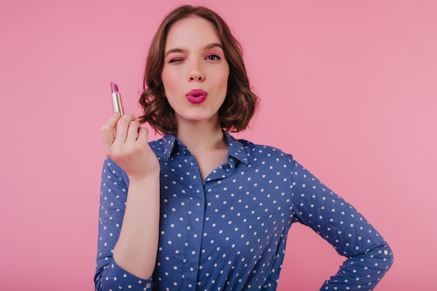 Atractiva mujer blanca con corte de pelo corto posando con expresión facial besos mientras se maquilla. Retrato interior de una agradable niña caucásica con lápiz labial.