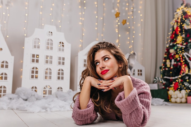 Atractiva mujer de cabello castaño con labios rojos y dulce sonrisa se encuentra en una habitación blanca decorada llena de juguetes navideños y un exuberante árbol de Navidad.