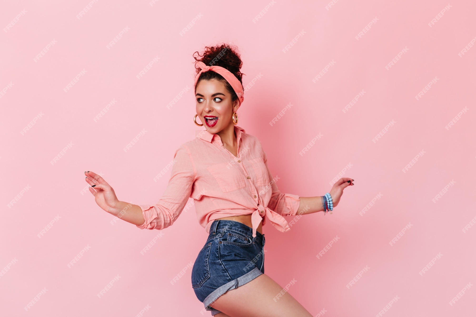 Atractiva mujer de cabello oscuro en ropa de estilo pin-up posando en el espacio rosa. retrato de dama con diadema y pantalones cortos de mezclilla. | Foto Gratis