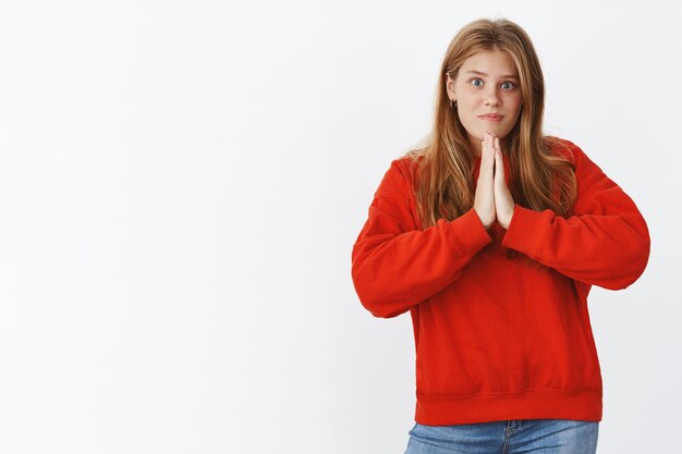 Atractiva mujer carismática joven amable y gentil pidiendo ayuda tomados de la mano en oración cerca del pecho y sonriendo linda con expresión tierna necesita favor sobre la pared gris