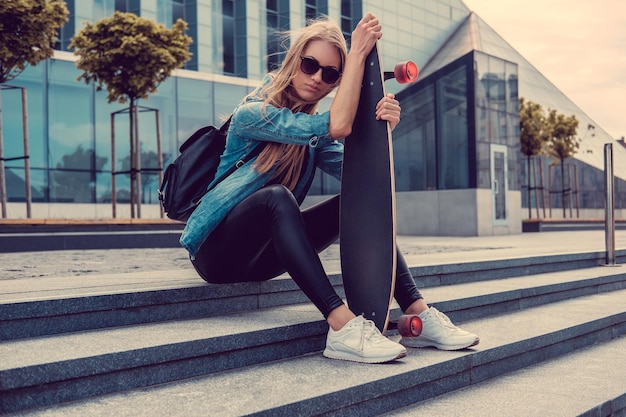 Atractiva mujer casual rubia se sienta en los escalones y sostiene Longboard.