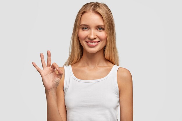 Atractiva mujer caucásica con cabello largo, expresión satisfecha, muestra bien, se siente feliz después de reunirse con un chico guapo, aislado sobre una pared blanca. Expresiones faciales humanas, lenguaje corporal.