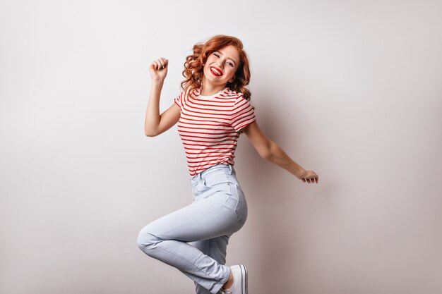 Atractiva mujer caucásica en camiseta a rayas divirtiéndose en la pared blanca. Chica de jengibre alegre de buen humor bailando.