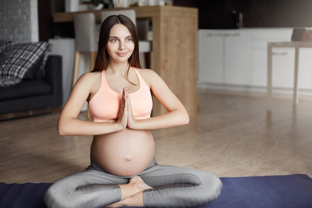 Atractiva mujer caucásica embarazada en ropa deportiva, sentada en pose de yoga en la almohadilla de rollo, sosteniendo las palmas juntas en gesto namaste, sonriendo a un lado mientras se relaja y piensa en el bebé