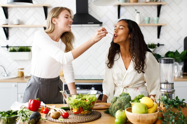 Atractiva mujer caucásica está alimentando a hermosa mujer mulata con ensalada fresca en la cocina de diseño moderno