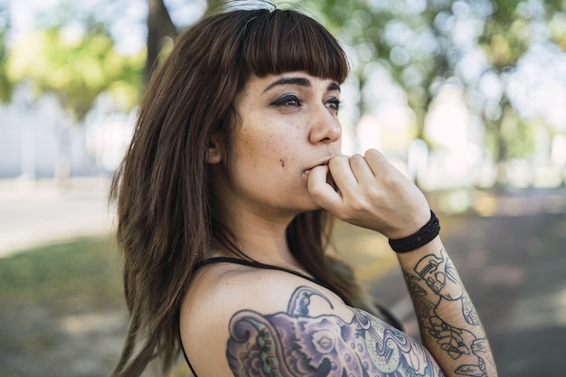 Foto gratuita atractiva mujer caucásica joven con tatuajes de pie en el parque y haciendo una linda cara