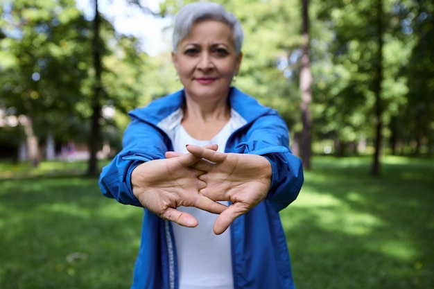 Atractiva mujer caucásica madura de pelo gris con chaqueta azul extendiendo los brazos y las manos, haciendo rutina de calentamiento mientras entrena al aire libre por la mañana. Enfoque selectivo en palmas