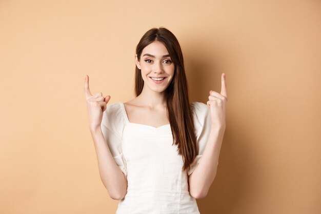 Atractiva mujer caucásica en vestido apuntando con el dedo hacia arriba sonriendo y mostrando banner promocional de pie sobre ...