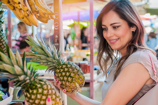 Atractiva mujer de compras en el mercado verde. Closeup retrato joven y bella mujer recogiendo, la elección de frutas, piñas. Expresión de la cara positiva emoción sensación de estilo de vida saludable